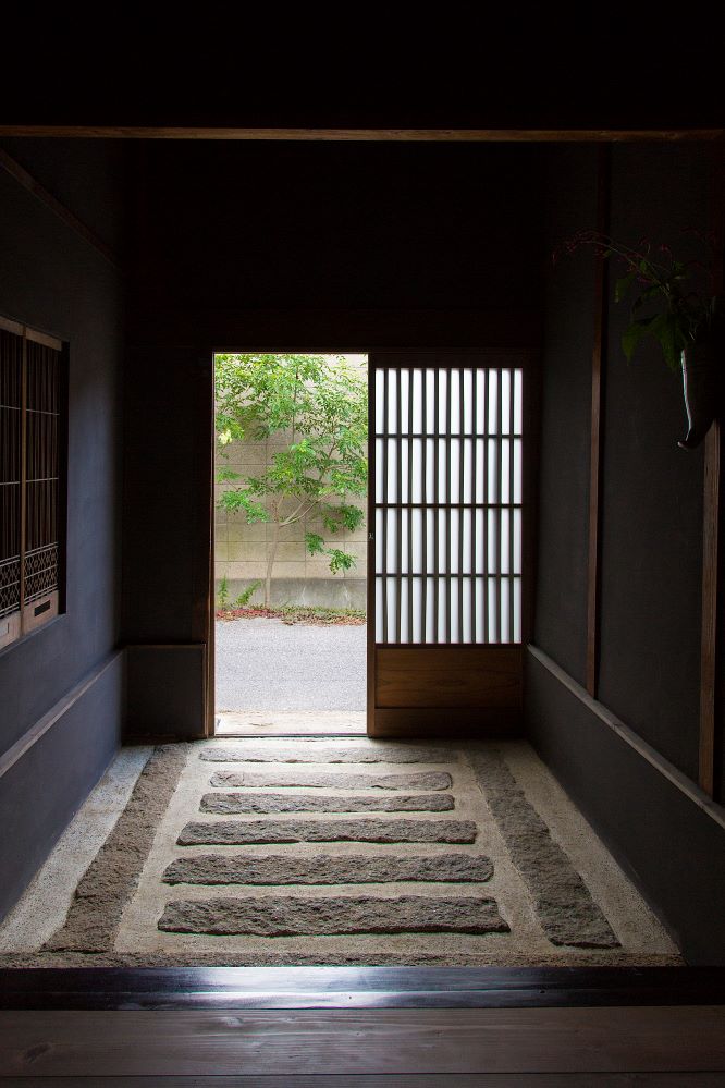 A serene corridor features textured stone flooring leading to an open door. Natural light enters from outside, illuminating a small garden visible beyond the door. Traditional sliding doors with vertical slats add a warm touch to the interior space.