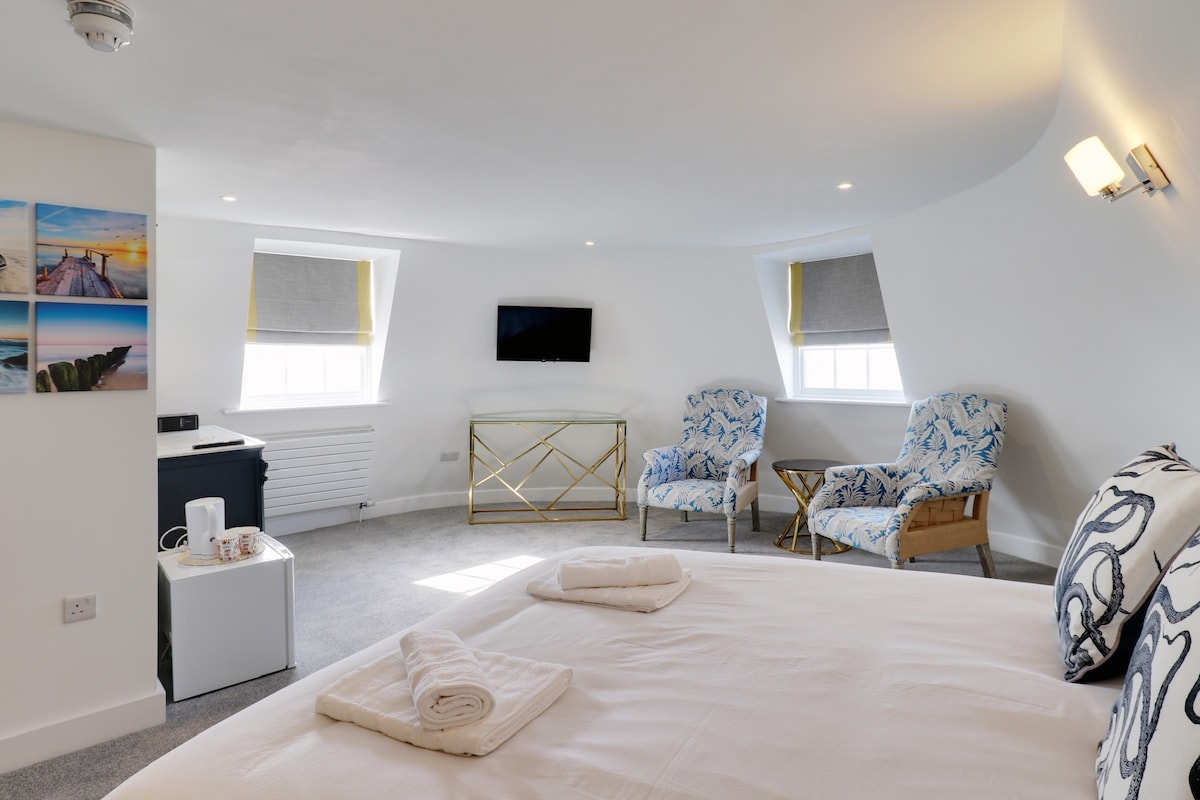 A spacious master suite features a large bed with neatly arranged towels. Two elegant chairs are positioned nearby, complemented by a small accent table. A wall-mounted television is visible, and a mini fridge is discreetly placed at the corner. Natural light filters through partially opened blinds.