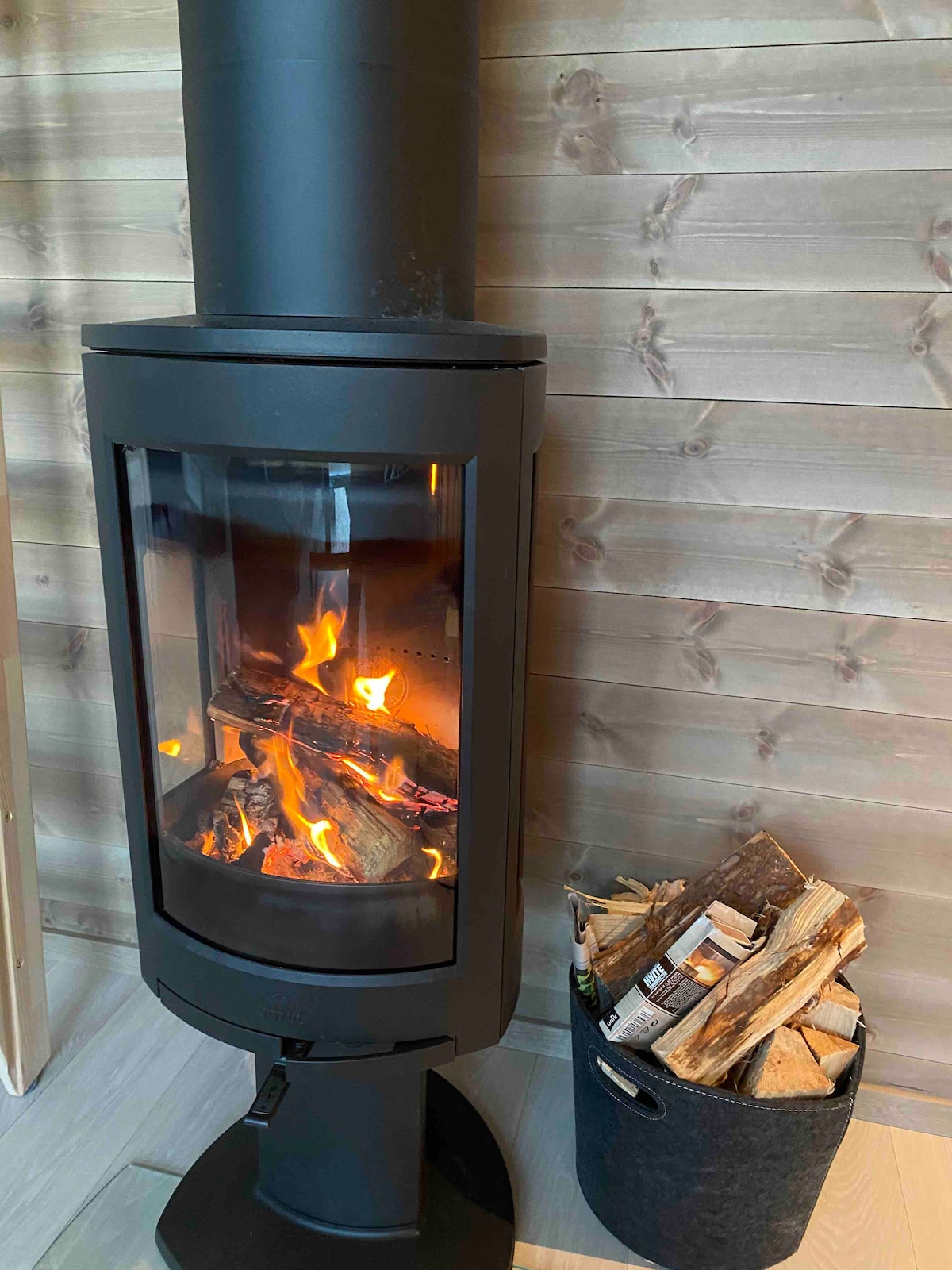A modern stove is positioned against a wooden panel wall, displaying a gentle flame through its glass panels. A nearby container holds neatly stacked firewood, providing easy access for fueling the fire.
