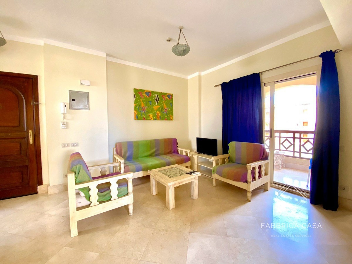 A spacious living area features a comfortable seating arrangement with colorful cushions on two sofas and a matching armchair. A light wooden coffee table is centered in front of the seating, complemented by a small television against the wall. Natural light enters through sliding glass doors, adorned with dark blue curtains.