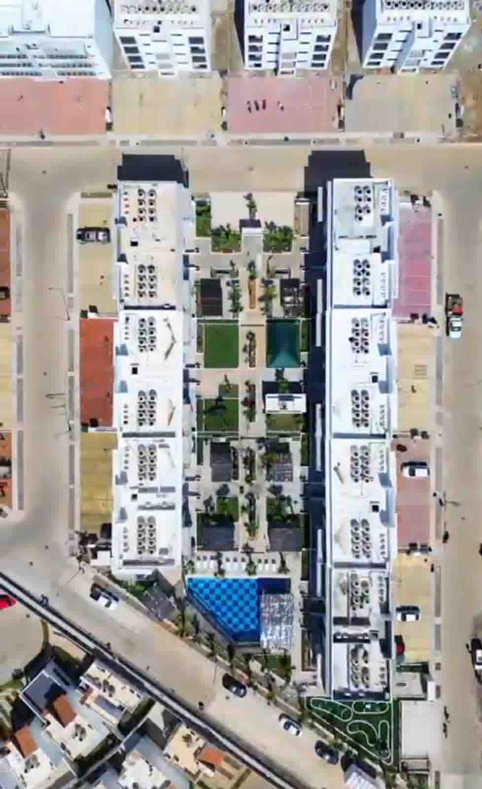 An aerial view displays a modern accommodation complex with multiple white buildings. Green spaces and walkways are seen interspersed among the structures. A blue section, possibly a pool, is visible in the center, surrounded by landscaped areas and pathways connecting different parts of the property.