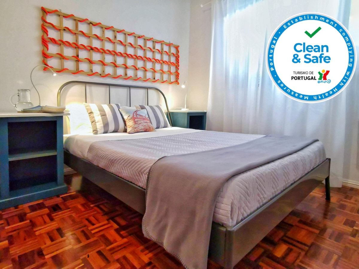 A well-lit room features a spacious bed with a light-colored duvet and decorative pillows. Unique wall art in bright colors complements the furnishings. Two bedside tables frame the bed, and natural light streams through a sheer curtain, enhancing the inviting ambiance.