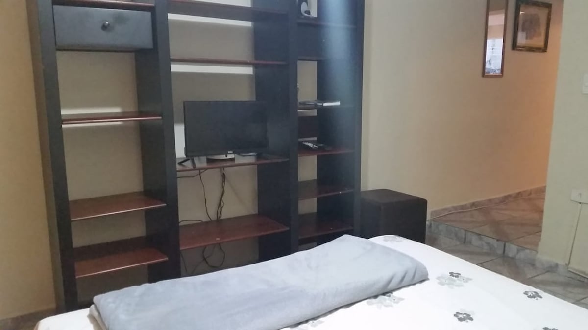 A cozy sleeping area is observed, featuring a spacious bed with a light grey blanket neatly placed. A compact television is situated on a black shelf unit, accompanied by an array of open storage spaces. The room is connected to a softly lit hallway.