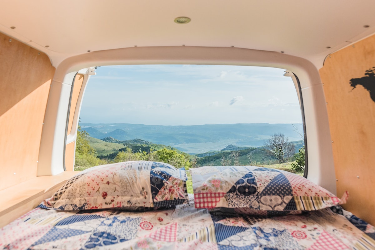 A scenic view is framed by the van's large rear window, showcasing rolling hills and distant mountains. The interior features a comfortable sleeping area with two pillows resting on a patterned duvet cover. Natural light fills the space, highlighting the wooden paneling.