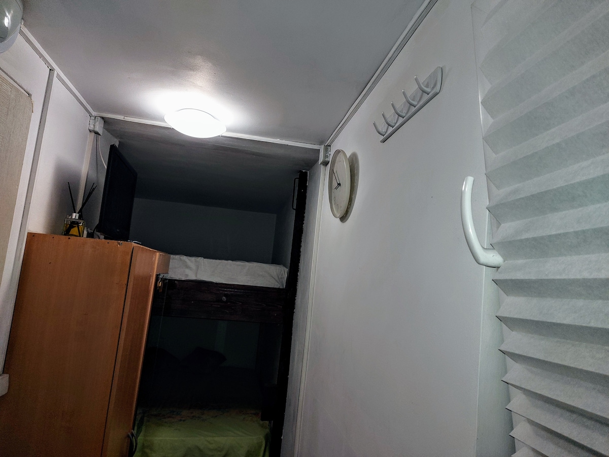 A wall-mounted light fixture illuminates the room, highlighting a wooden bunk bed with a lower sleeping area. A simple wall clock and hooks provide functional elements against the light-colored walls. Nearby, a wooden cabinet offers storage, contributing to the organized space.