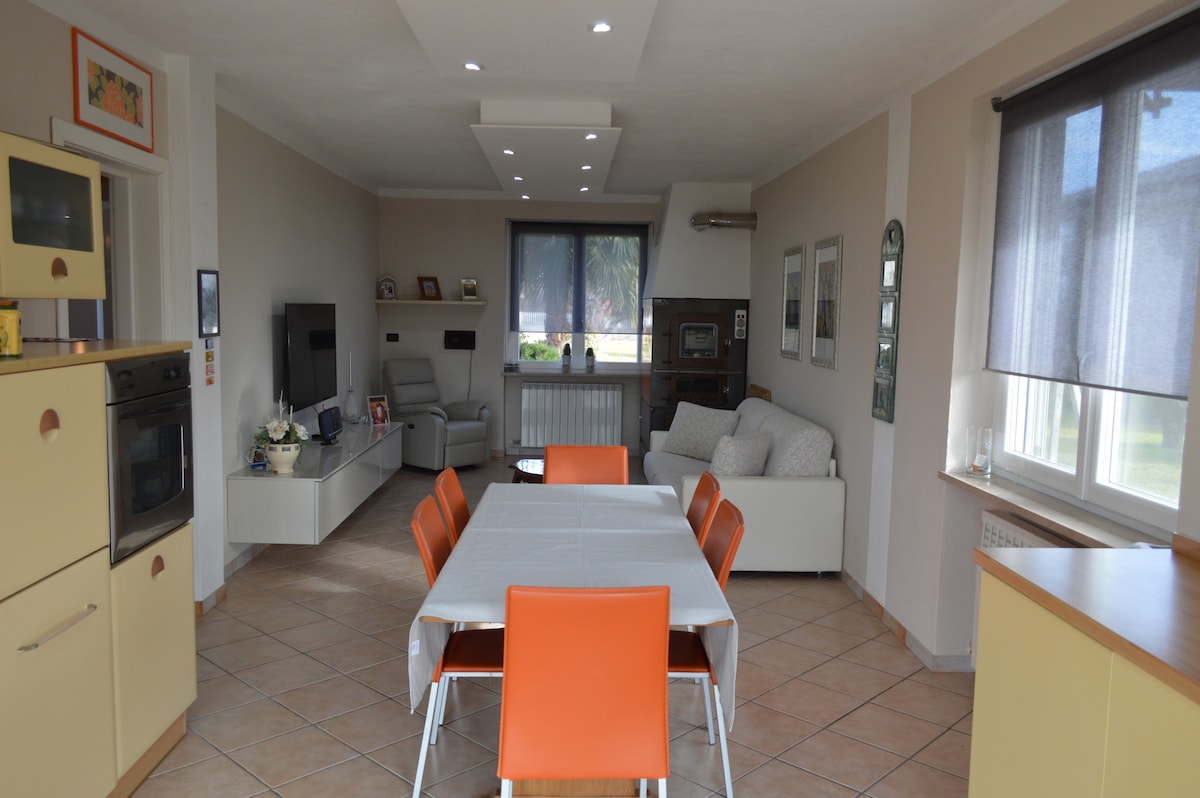The spacious living area incorporates a dining table with orange chairs and an equipped kitchen. A comfortable sofa and TV are present in the adjoining lounge space. Natural light filters through large windows, and neutral tones create a calming environment.
