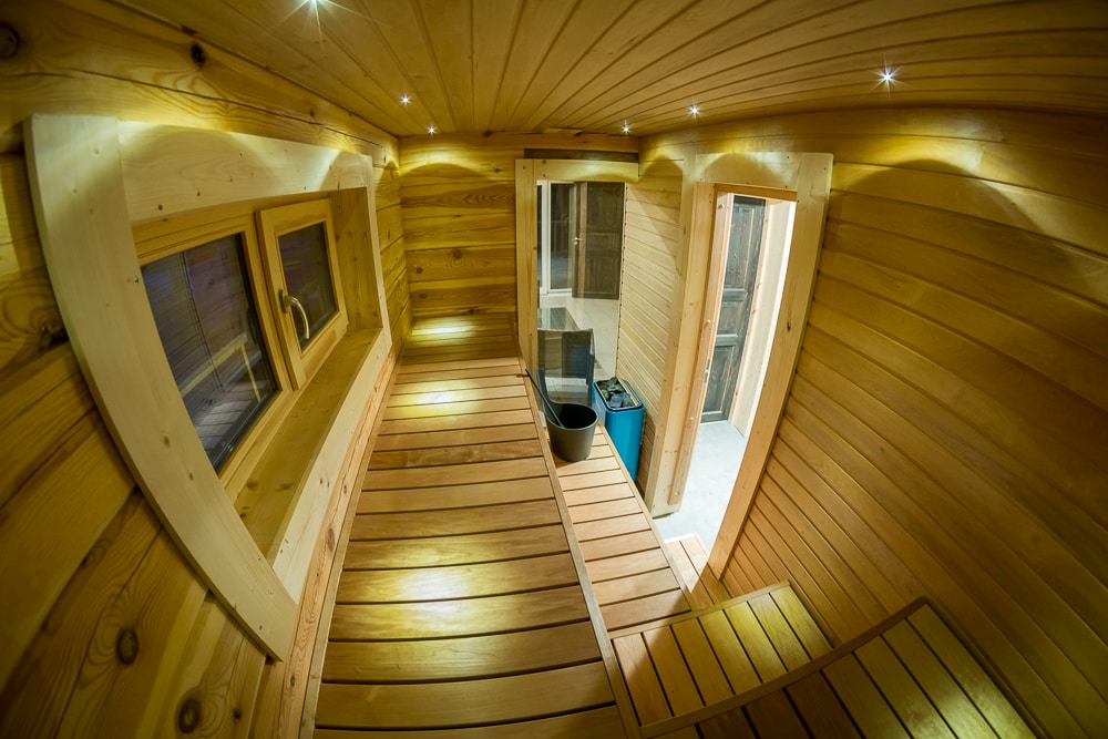 A well-crafted wooden sauna features warm wooden benches and walls, illuminated by recessed lighting. Windows allow for natural light while wooden slats create a soothing ambiance. An entryway leads to a small area with a bucket and additional space visible beyond.