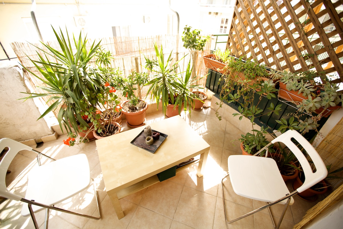 A small balcony area is furnished with a coffee table and two chairs, surrounded by leafy potted plants. Natural light brightens the space, creating a refreshing atmosphere. A trellis in the background adds privacy while enhancing the setting with greenery.