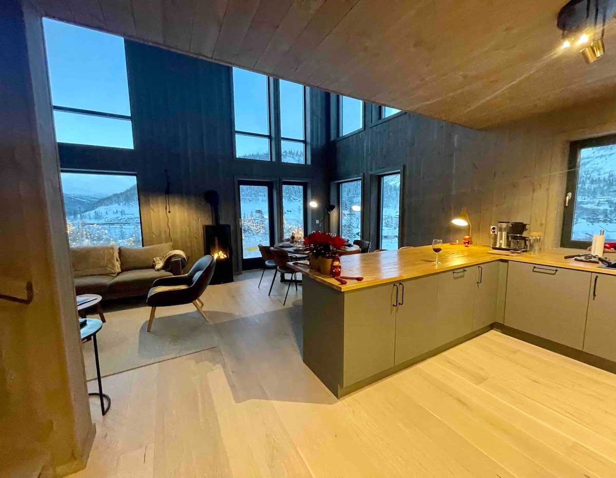 An open living space is highlighted by large windows that frame panoramic views of the surrounding mountains. A modern kitchen island contrasts with the natural wood finishes, and comfortable seating invites relaxation in the cozy lounge area.