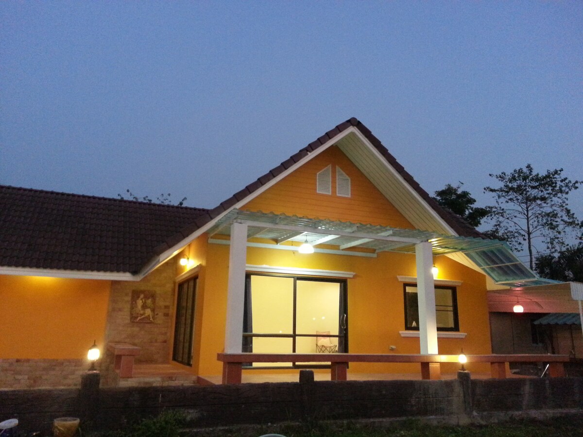 A charming orange house is presented, featuring a sloped roof and a covered porch. Bright lights enhance the exterior as evening approaches. Large glass doors lead into the interior, with ample outdoor space visible for relaxation or social gatherings.