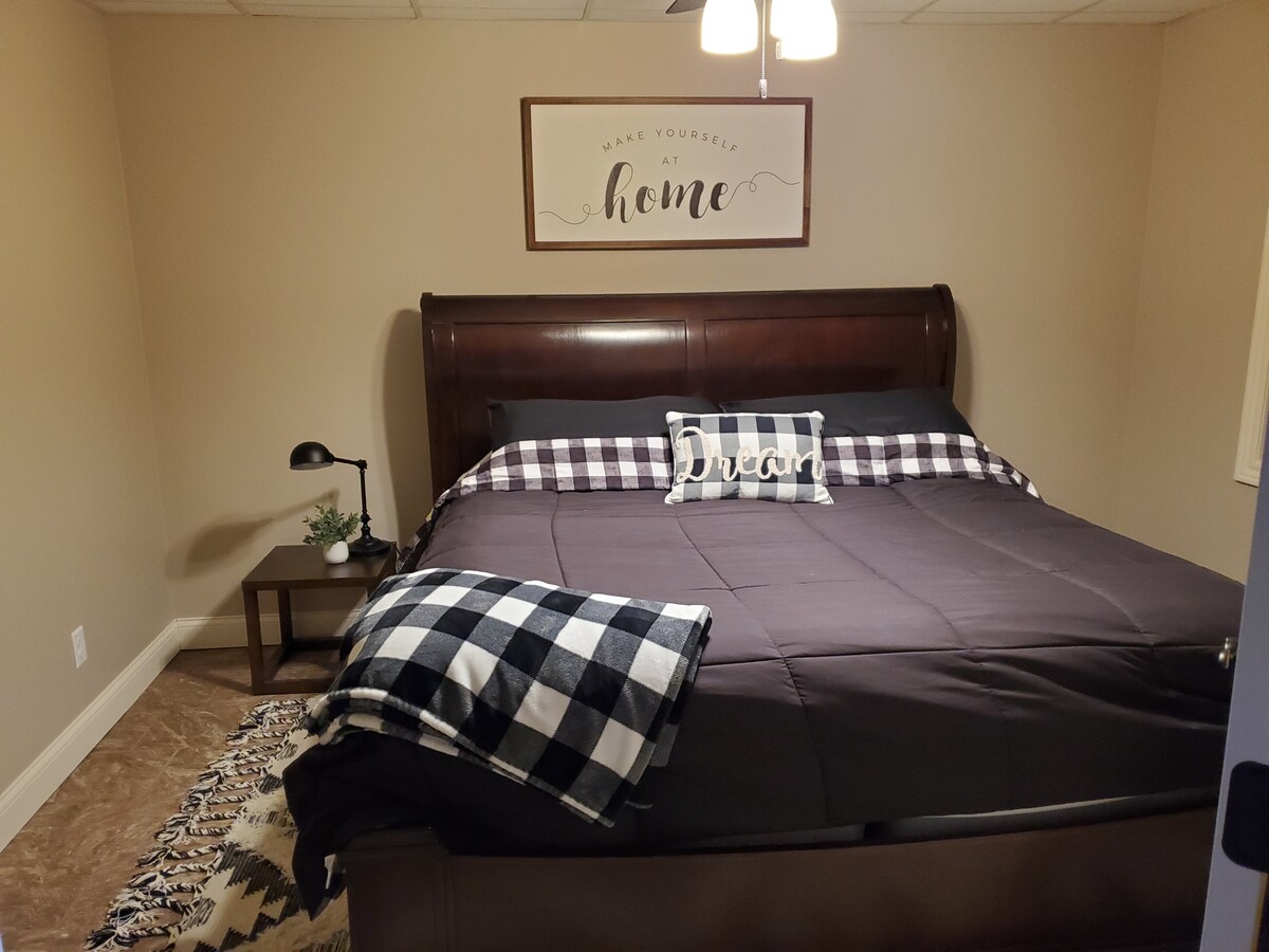 A spacious bedroom features a large, dark wood bed adorned with a dark blanket and plaid pillows. A bedside table with a lamp is positioned next to the bed, and a small plant adds a touch of greenery. A decorative wall sign emphasizes a welcoming message.