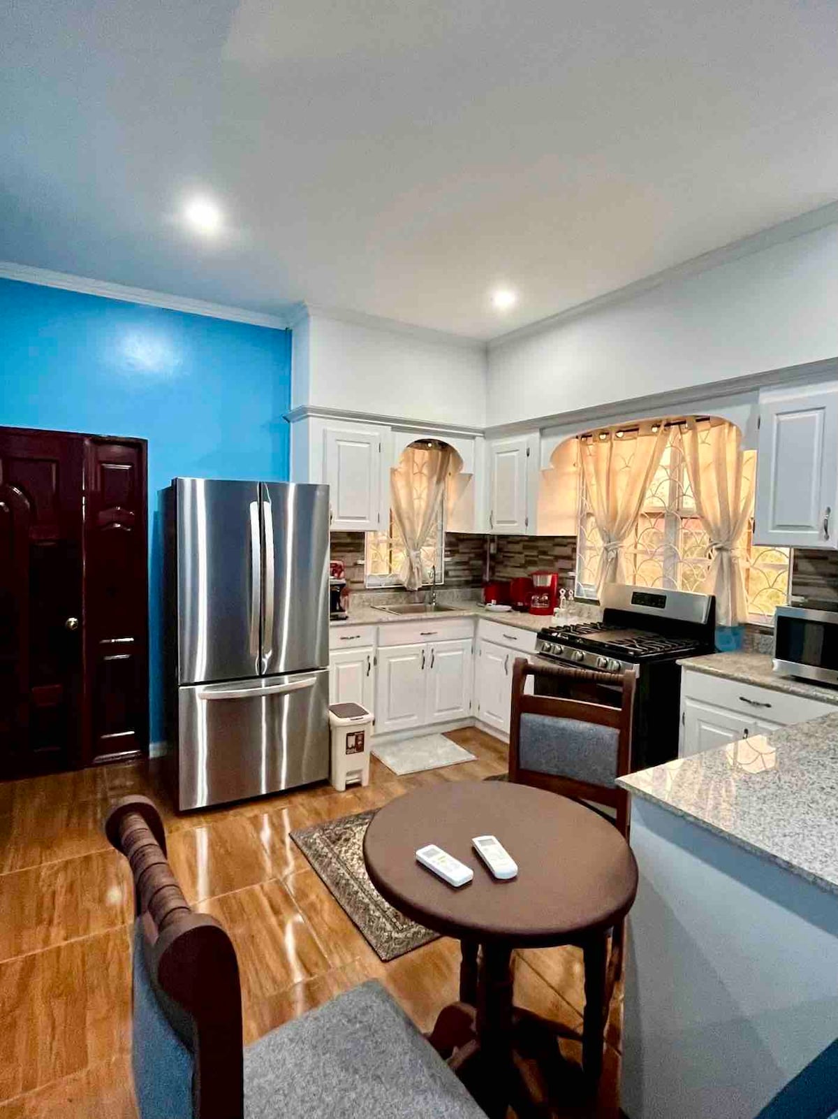 A modern kitchen is equipped with new stainless-steel appliances, including a refrigerator and stove. Cabinets in white contrast with the granite countertops. Soft yellow curtains frame the windows, and a small round table with two chairs is positioned nearby. The walls are painted in a light blue hue.