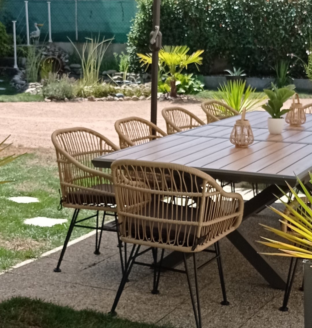 A spacious outdoor dining area is presented with a stylish table surrounded by several woven chairs. Greenery and small plants adorn the setting, enhancing the tranquil garden atmosphere. A parasol provides shade, creating a comfortable space for relaxation and dining.
