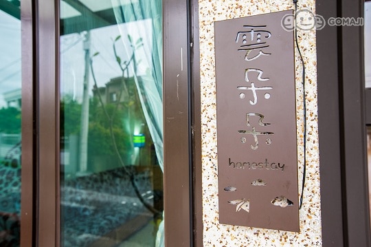 A sign displaying the name of the homestay is mounted on a stone-textured wall. The sign's letters are cut out, allowing light to filter through and create an inviting presence. Soft curtains dance gently in the breeze, adding a touch of warmth to the entrance.