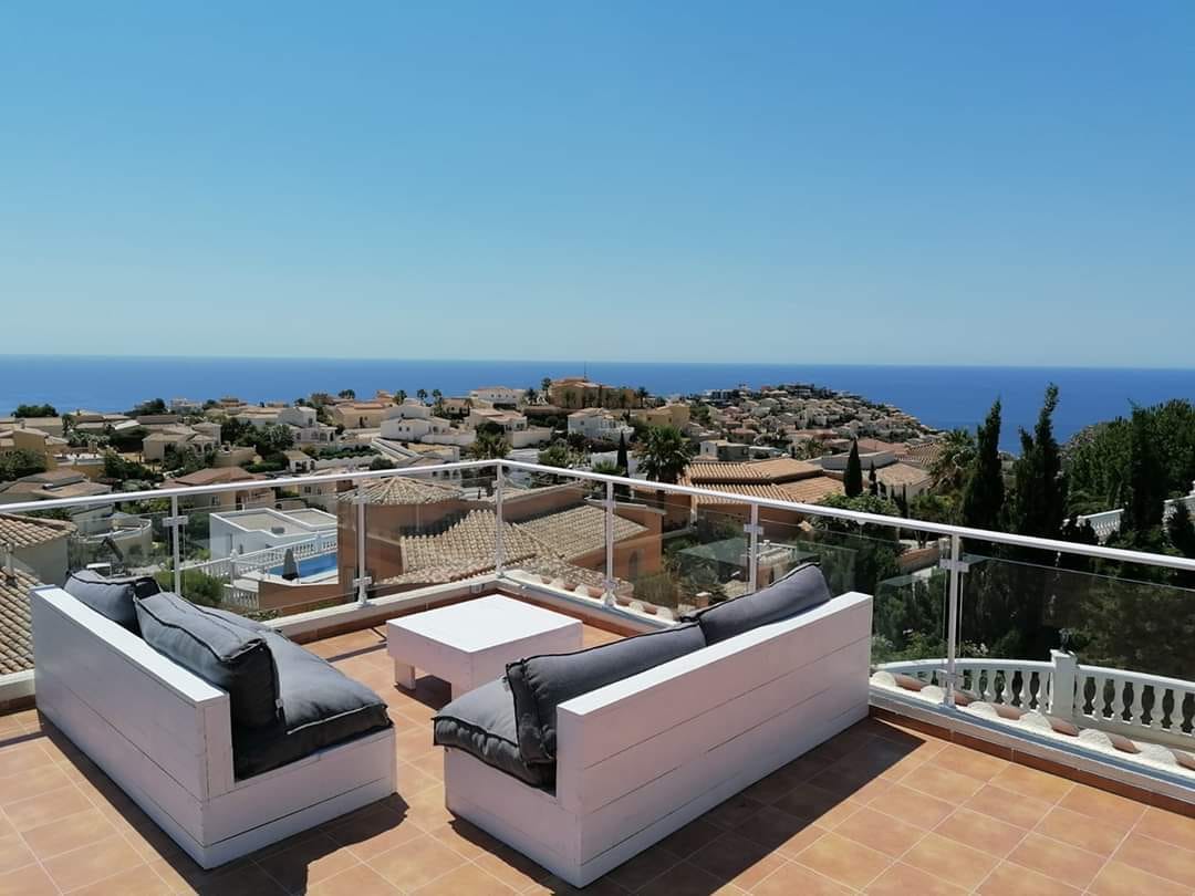 A spacious terrace offers a panoramic view of the ocean, framed by a glass railing. Two cushioned lounge chairs and a coffee table are arranged for relaxation, with the surrounding landscape featuring residential rooftops and greenery under a clear blue sky.