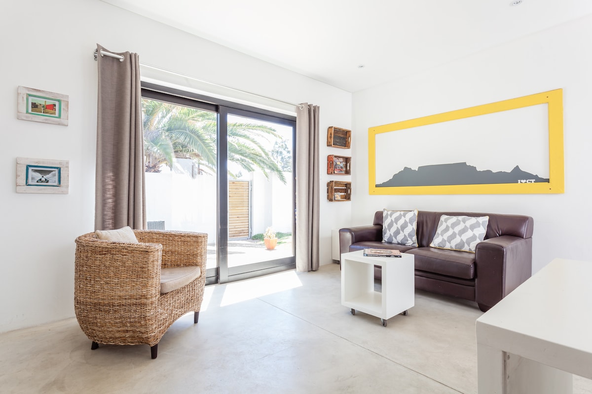 A cozy lounge area features a brown leather sofa and a woven armchair. A small white table is positioned in the center. Large glass doors provide access to an outdoor space, while a vibrant yellow artwork depicting Table Mountain is displayed on the wall.