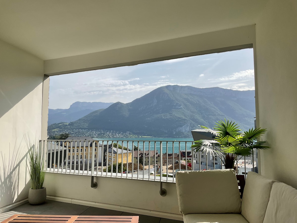 A spacious loggia offers stunning lake views framed by the surrounding mountains. Comfortable seating is provided, complemented by a small wooden table. Natural light illuminates the area, enhancing the greenery of potted plants positioned nearby.