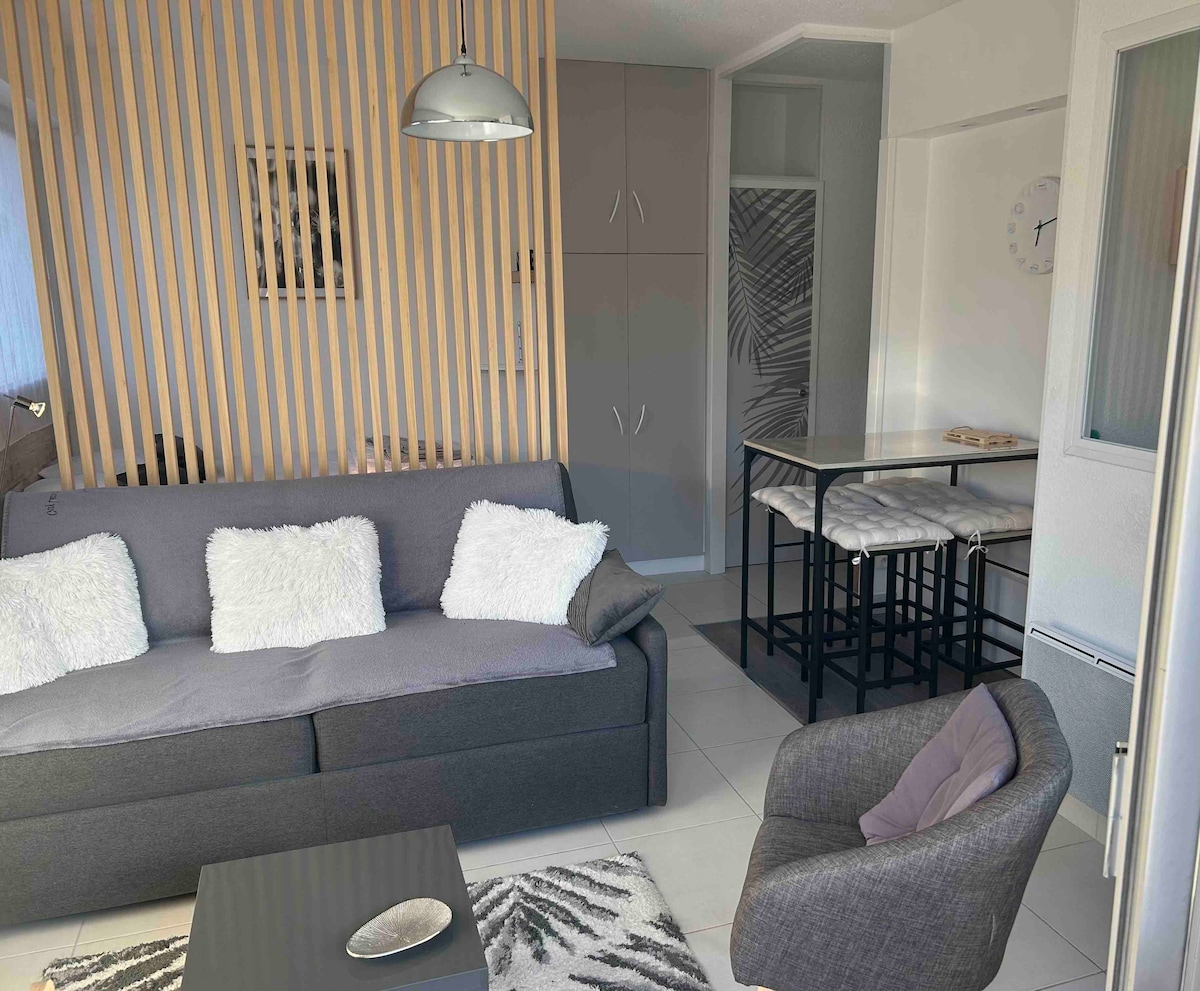A cozy living area features a gray sofa adorned with white cushions. A sleek coffee table sits on the floor, while a high table with stools is positioned nearby. Light wood slats provide a touch of warmth, and natural light fills the space.