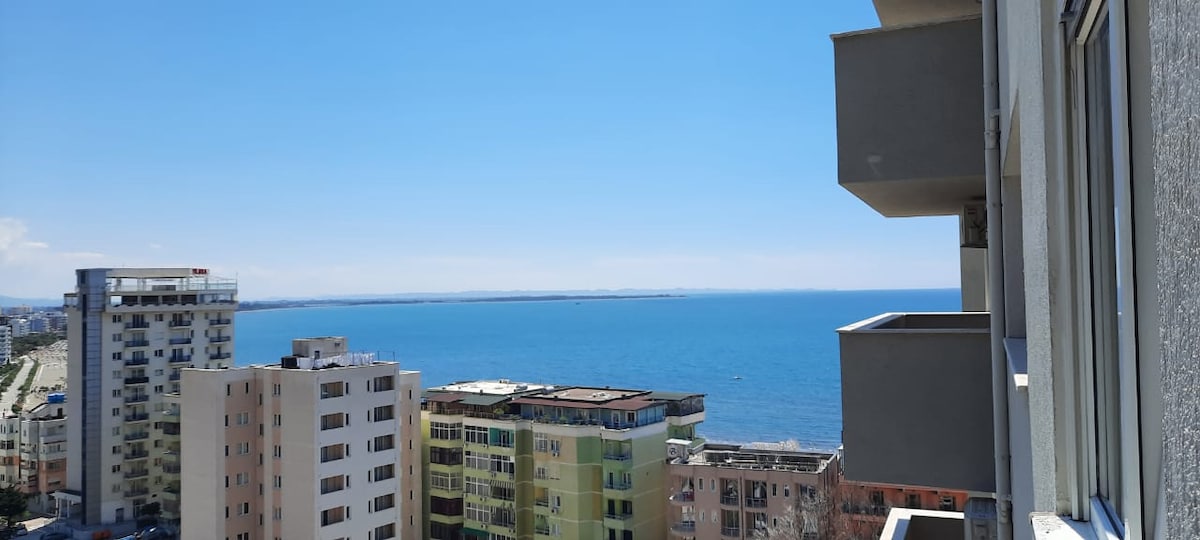 A scenic view is captured from a height, showcasing a vibrant blue sea meeting the sky. Coastal buildings in various colors are lined along the shoreline, with the calm waters reflecting the clear sky.