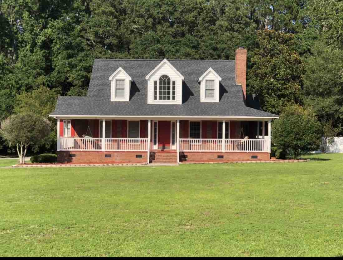 A spacious country home is surrounded by lush greenery and sits on a grassy expanse. The structure features a wrap-around porch supported by white columns, with decorative red shutters accentuating the windows. A brick chimney rises from the roof, completing the inviting façade.
