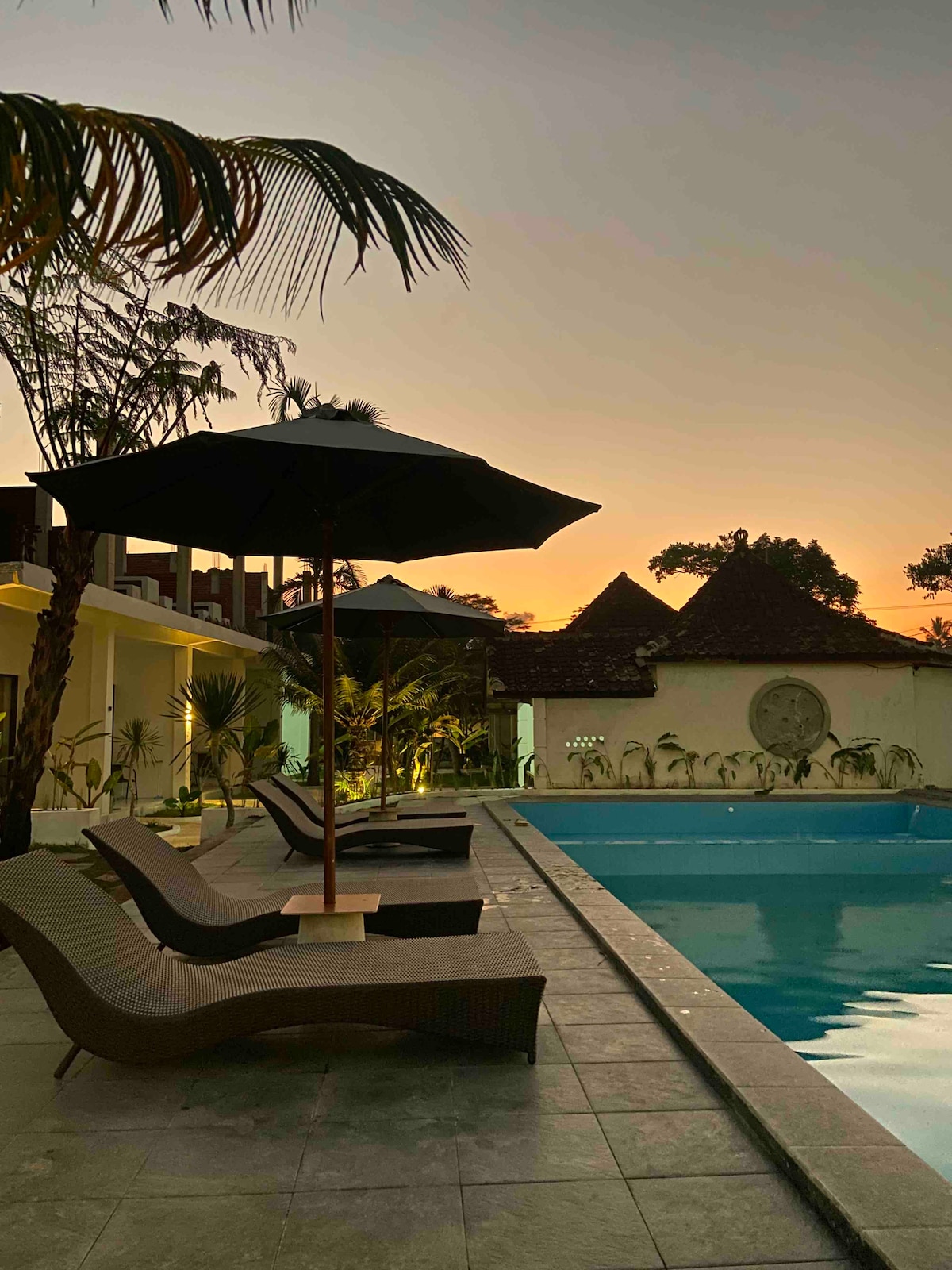 A tranquil outdoor area features a swimming pool lined with loungers under large umbrellas. Palm trees sway gently nearby, and the scene is bathed in the warm colors of sunset, creating a serene atmosphere.