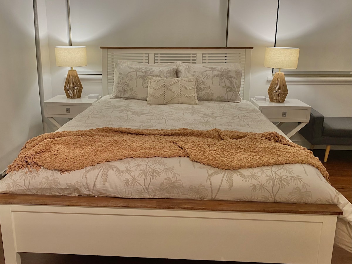 A spacious bed with a neutral-toned floral pattern is centered in the room, flanked by two bedside tables featuring lamps that emit a soft glow. A textured throw blanket is casually draped over the bed, adding warmth to the inviting atmosphere.