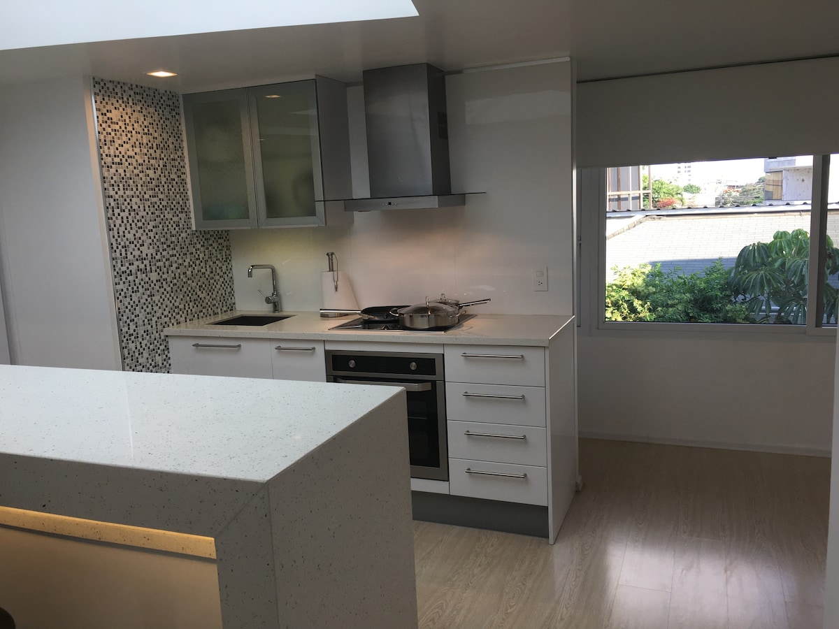 A modern kitchen features sleek white cabinetry and a contrasting mosaic tile backsplash. Stainless steel appliances, including a stove and oven, are visible among the prep areas. A window allows natural light to illuminate the space, enhancing the open feel.