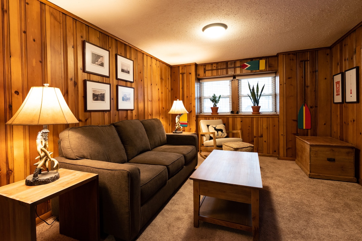 The living room features warm pine walls, offering a cozy atmosphere. A dark, spacious sofa is positioned alongside a wooden coffee table. Two lamps illuminate the space, framed artwork adorns the walls, and potted plants are placed on the windowsill, adding a touch of greenery.