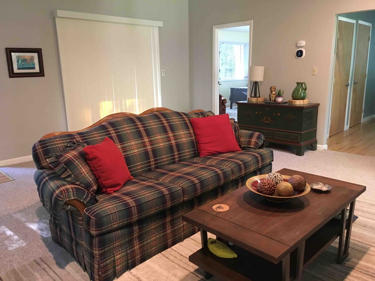 A spacious living area features a plaid sofa with red accent pillows, positioned next to a wooden coffee table. Natural light enters through nearby windows, illuminating the soft carpeting and neutral walls that create a comfortable environment.