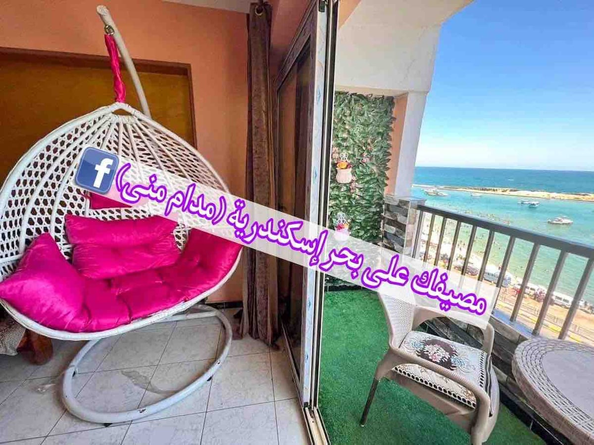 A spacious balcony features a hanging chair, offering sweeping views of the sea. The vibrant colors of the outdoor area complement the inviting seating space. The wall near the balcony is adorned with greenery, enhancing the overall atmosphere.