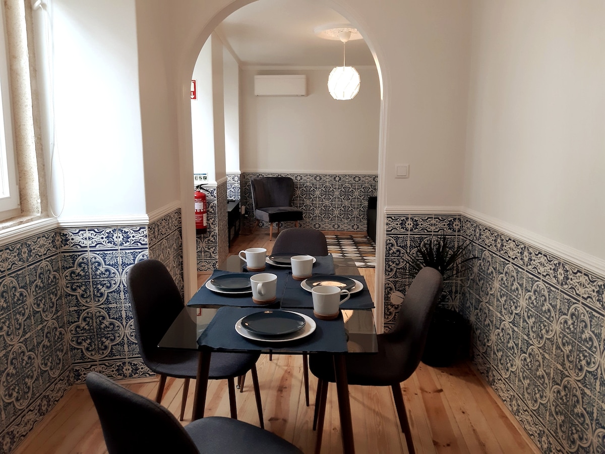 A dining area is displayed with a glass table surrounded by four modern gray chairs. The table is set with white dishware and blue placemats. Decorative blue tiles adorn the lower walls, while natural light fills the space through nearby windows.