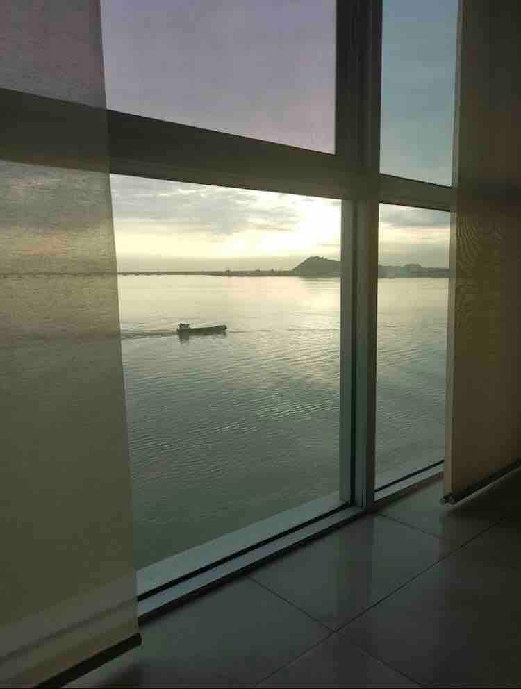 Large floor-to-ceiling windows provide a panoramic view of the Guayas River, showcasing calm waters and distant hills. A small boat can be seen gliding across the river's surface, reflecting the tranquility of the scene during the early morning or late afternoon light.