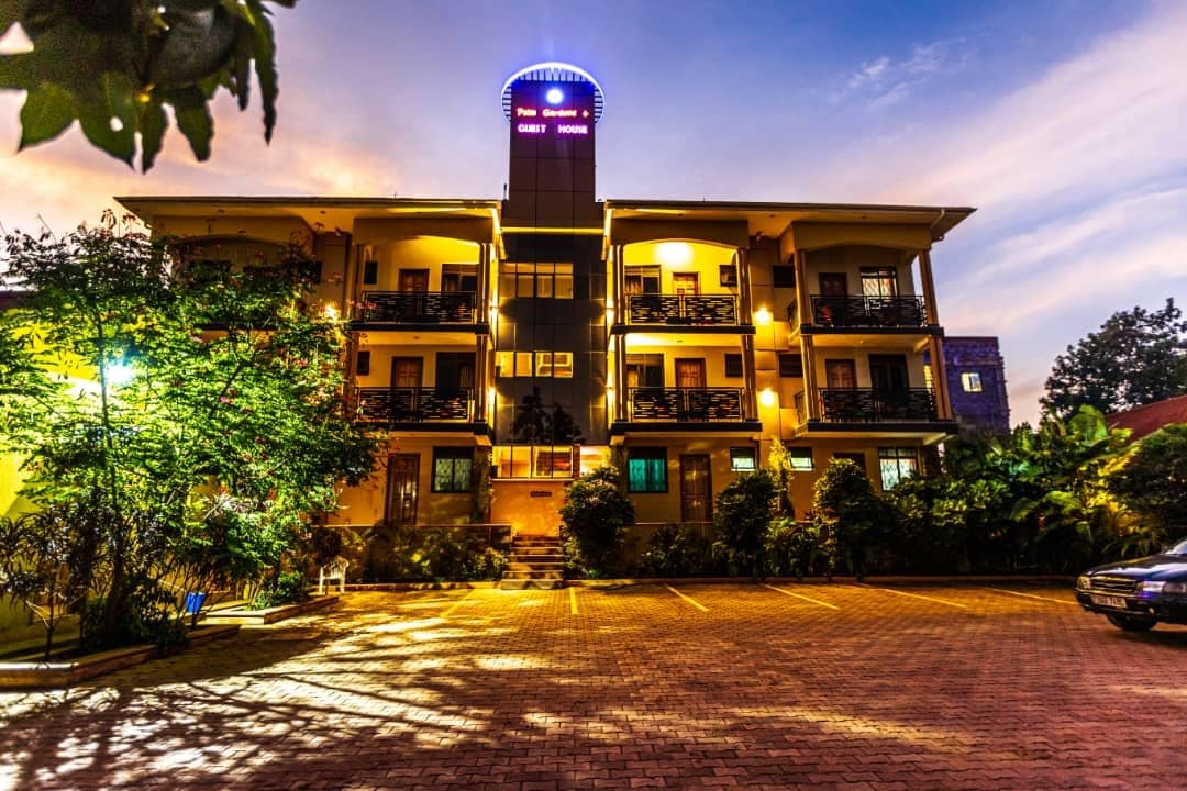 An inviting three-story building is illuminated by outdoor lighting, showcasing balconies on the upper levels. Lush greenery surrounds the exterior, while well-maintained parking spaces are visible in the foreground against a twilight sky.