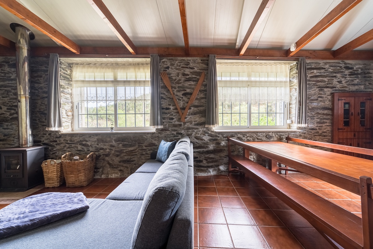 A spacious lounge area features a gray sectional sofa and a large wooden dining table. Natural light enters through two windows adorned with light curtains, illuminating the textured stone walls. A wood-burning stove adds warmth to the environment, with wicker baskets placed nearby.