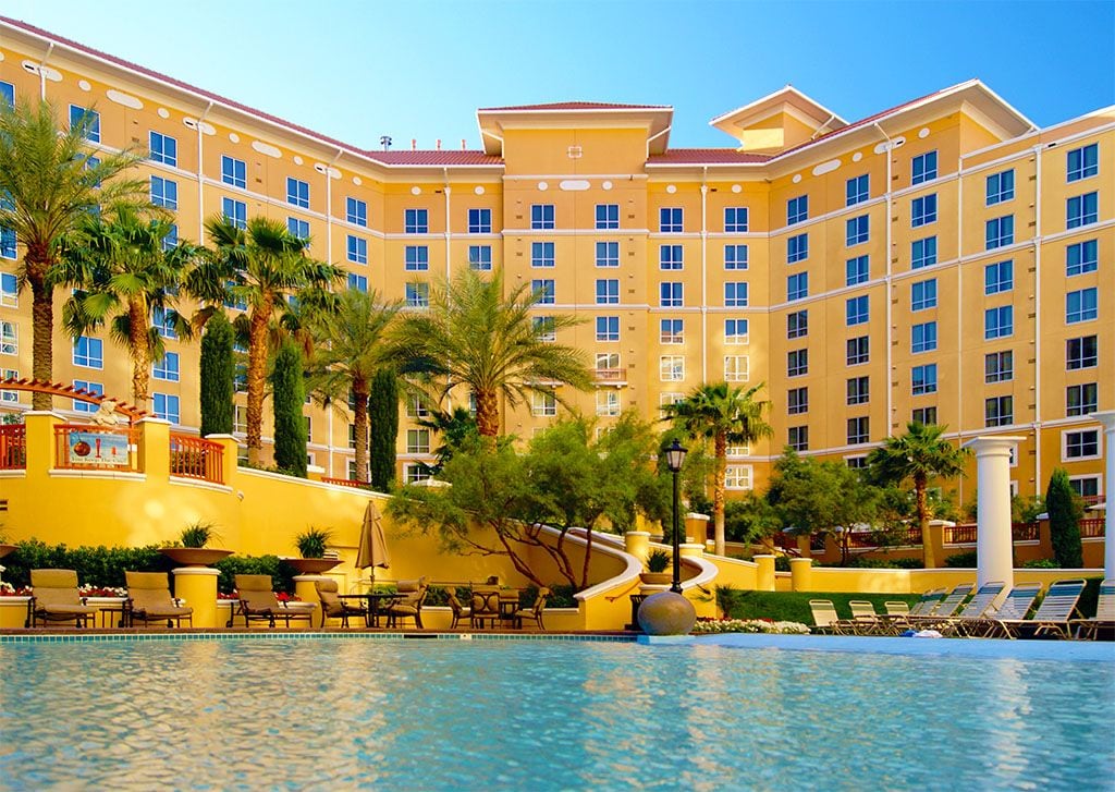 A vibrant yellow resort is showcased in the background, framed by lush palm trees and greenery. The foreground features a glistening pool with lounge chairs and umbrellas for relaxation under the sun, inviting guests to enjoy the outdoor space.