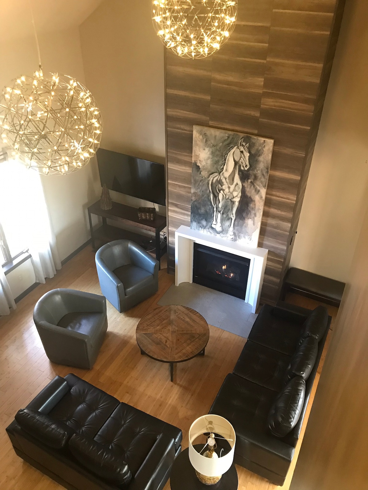 The living room presents a modern design featuring a fireplace framed by textured wall panels. A circular glass coffee table is positioned at the center, surrounded by two gray armchairs and a leather sofa. A piece of horse-themed artwork hangs prominently on the wall, complemented by spherical light fixtures.