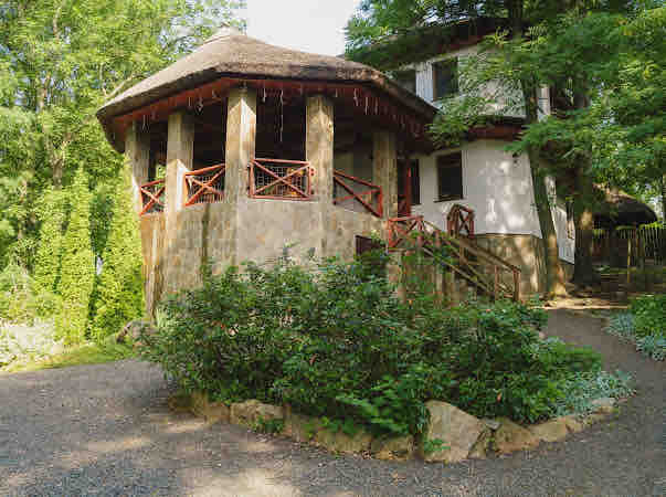 A round structure with a thatched roof is surrounded by lush greenery and stone pathways. The stone facade features large, open balconies with wooden railings, providing access to the outdoor areas. Mature trees and shrubs create a serene atmosphere around the entryway.