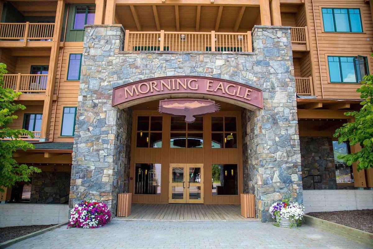 The entrance of Morning Eagle Lodge is framed by a stone façade, featuring a large wooden sign with the name 'MORNING EAGLE'. Colorful flower beds in front add vibrancy, while the spacious wooden deck welcomes guests into the building.