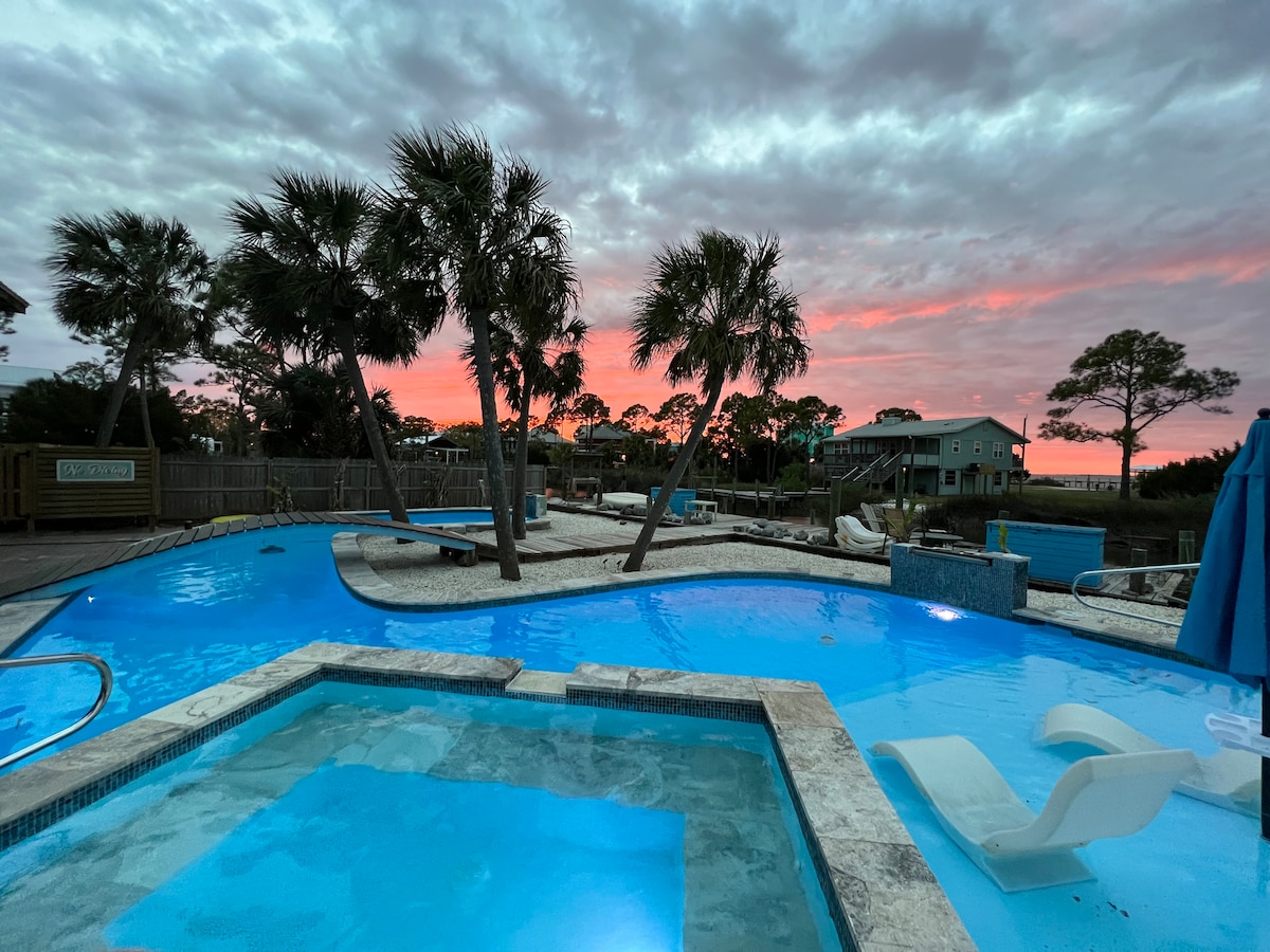 A serene saltwater pool is highlighted, featuring a hot tub and lounge chairs. Palm trees frame the area, and a colorful sunset casts reflections on the water, while neighboring buildings are subtly visible in the background.