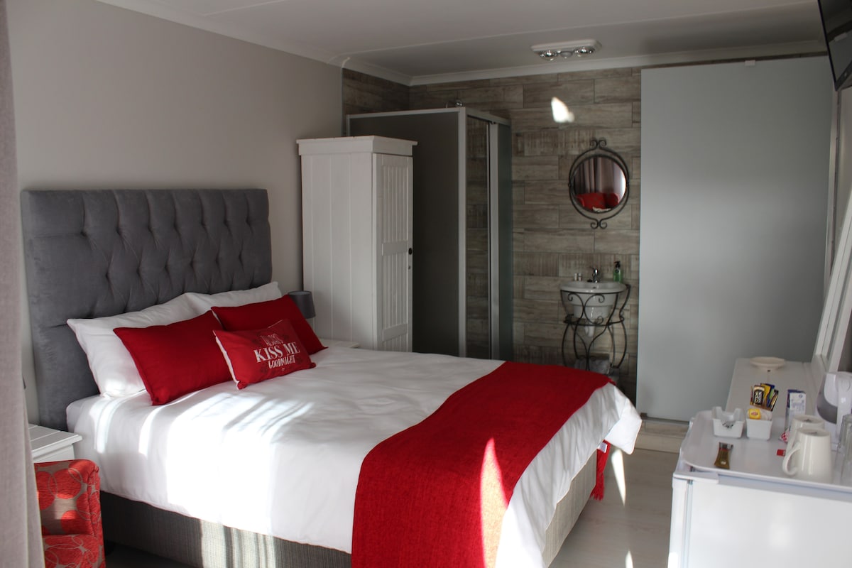 A well-appointed bedroom features a spacious bed dressed in crisp white linens, complemented by a bright red throw. A decorative mirror and a small side table are visible, while a wardrobe and a partitioned area provide additional storage options.