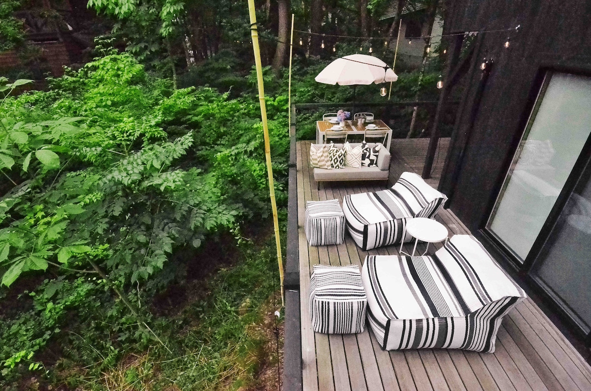 A spacious wooden deck features comfortable seating with striped cushions, complemented by a small round table. A parasol provides shade, while string lights add a subtle ambiance. Lush greenery surrounds the area, creating a serene outdoor setting for relaxation.