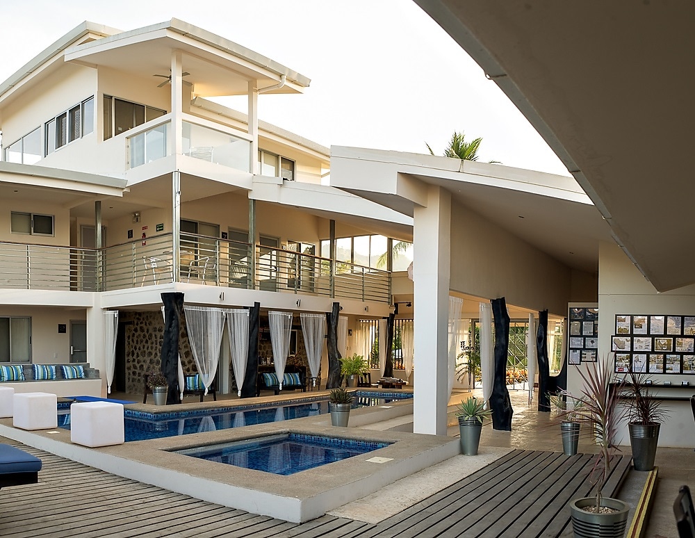 An expansive outdoor area is featured, showcasing a multi-level structure with smooth lines and large windows. A rectangular pool occupies the center, surrounded by shaded seating areas. Potted plants add a touch of greenery to the space, enhancing the tranquil environment.