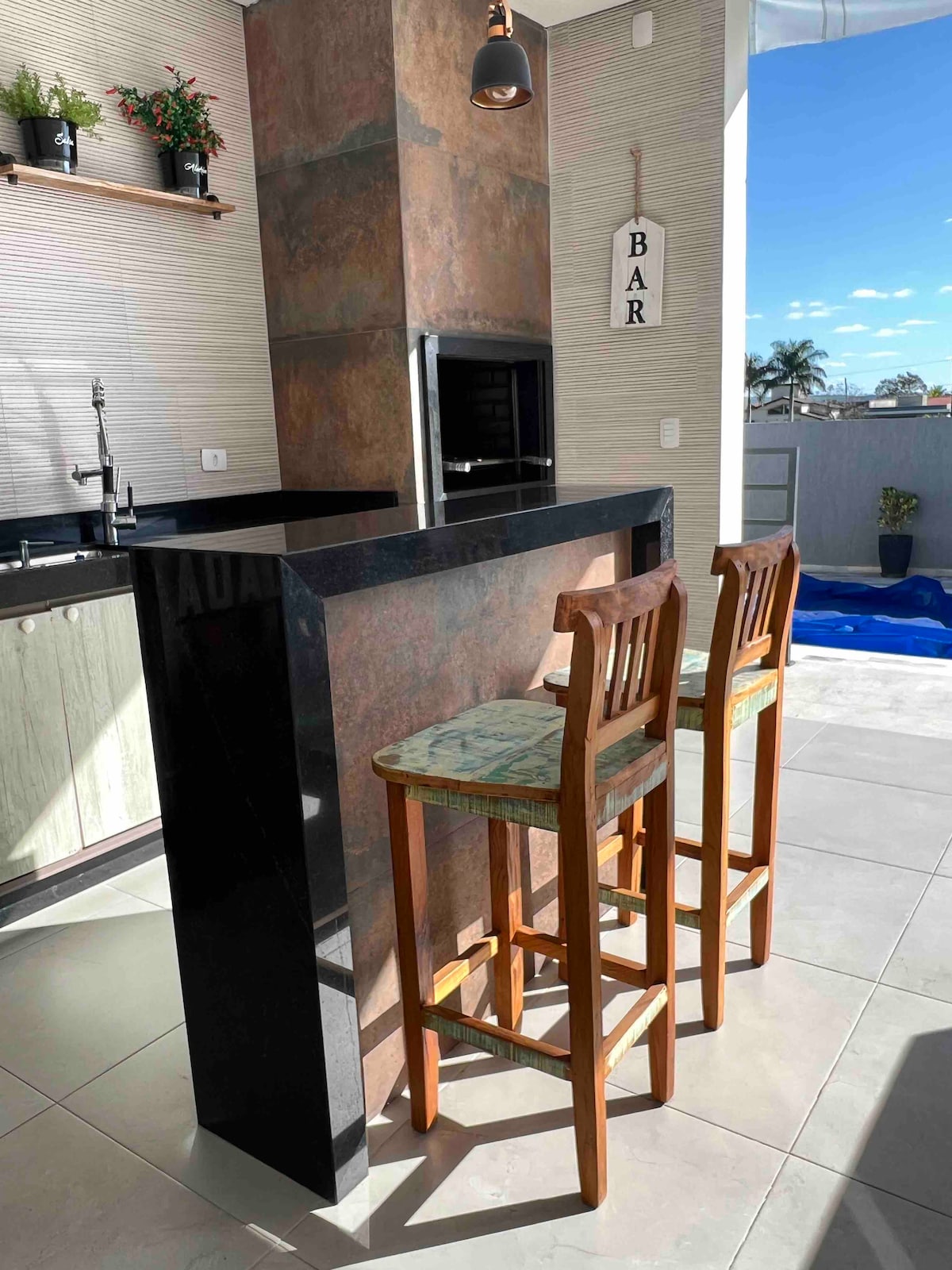 A modern kitchen area features a sleek black countertop with a grill and two wooden bar stools. Natural light illuminates the space, highlighting the contrasting textures of the dark surfaces and light cabinetry. Decorative plants are positioned on a shelf above the counter.