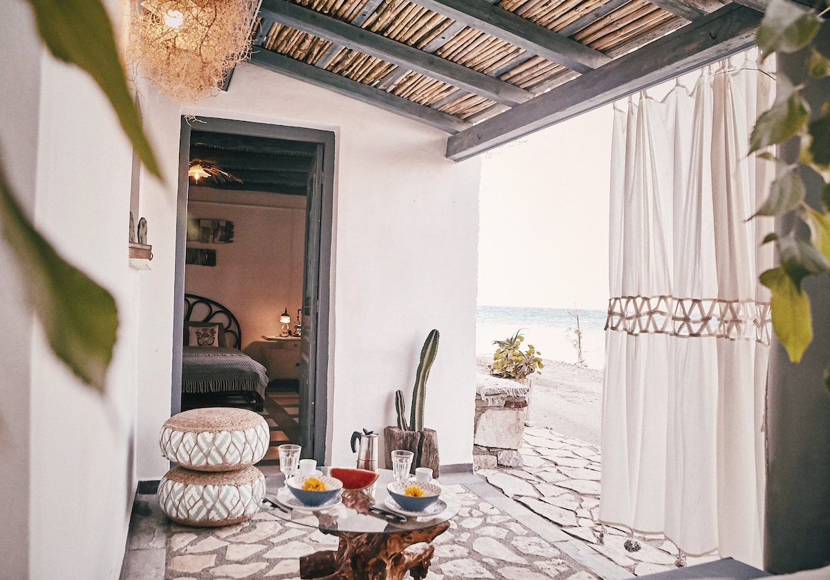 A covered patio showcases outdoor furniture with a view of the beach. Natural light enters through sheer curtains, highlighting stone flooring and decorative elements. In the background, a doorway leads into the bedroom, enhancing the sense of inviting space.