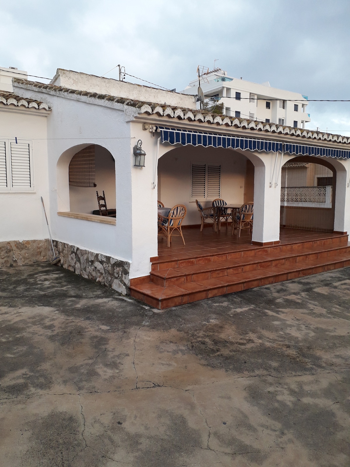 An outdoor terrace area features several wooden chairs positioned around a table. The steps lead up to the covered space, which is defined by archways and decorative tiles. The surrounding walls are painted white, contributing to a relaxed, open environment.