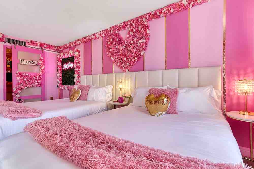 A brightly decorated room features two spacious beds adorned with plush white linens and shimmering pink accents. Pink flower arrangements are arranged on the walls, creating a heart shape. Soft lighting from bedside lamps enhances the cheerful atmosphere throughout the space.
