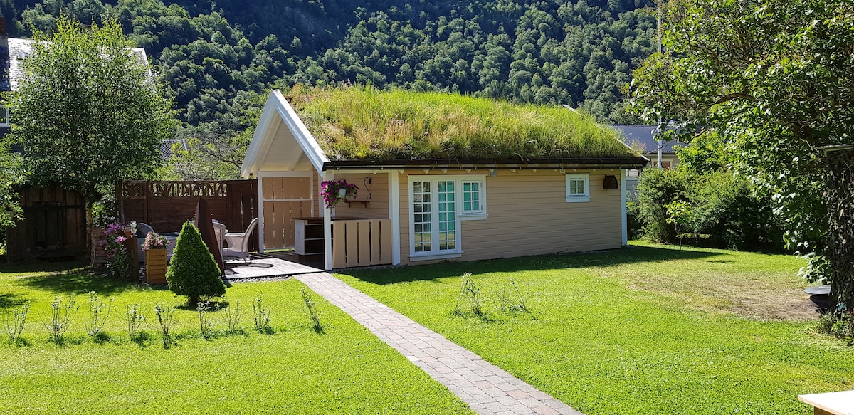 A charming cabin is set in a green garden, featuring a grassy roof and a well-maintained lawn. Outdoor seating is arranged on a stone patio, surrounded by flowering plants. The front facade includes large windows and wooden details, blending into the serene landscape.