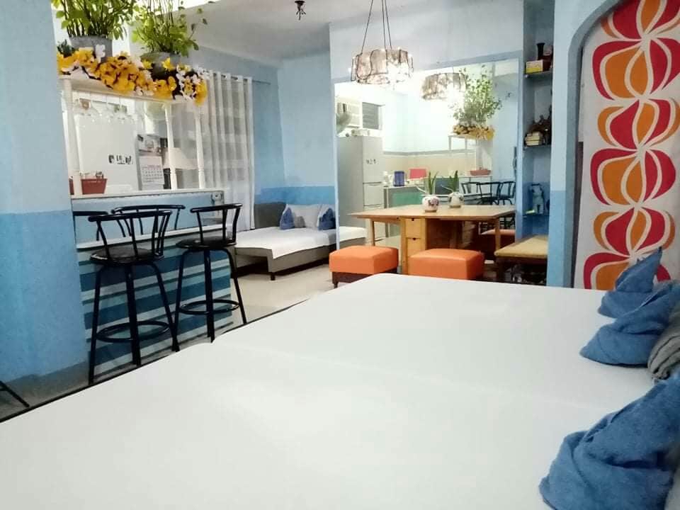 An inviting living space features a light blue color scheme with comfortable seating in the background. The room includes a spacious area with a large table and chairs, along with a breakfast bar and a small kitchenette. Natural light filters through the window, enhancing the welcoming atmosphere.