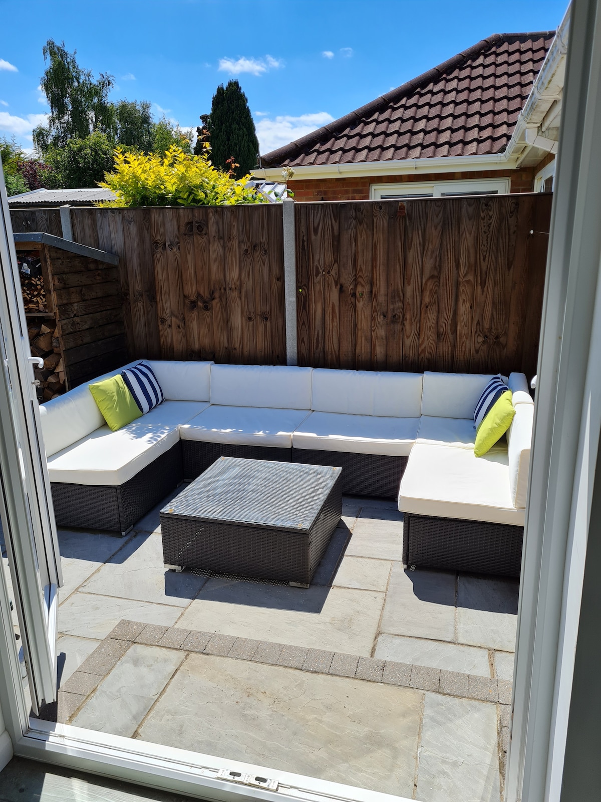 A private patio area features a modular outdoor seating arrangement with light-colored cushions and green accent pillows. A rectangular coffee table sits in the center, surrounded by a stone floor. The space is enclosed by wooden fencing and lush greenery in the background.