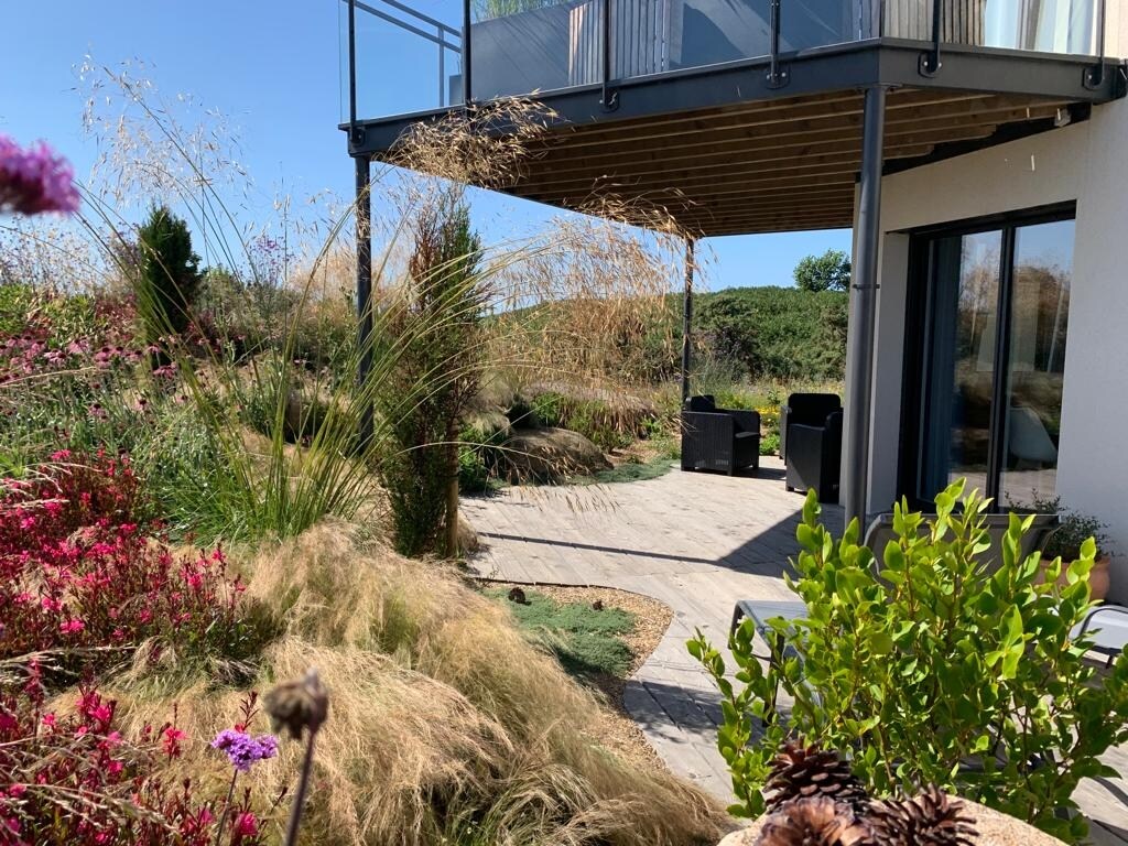 A spacious terrace extends from the studio, overlooking a beautifully landscaped garden filled with wild grasses and flowering plants. Comfortable seating areas are visible, inviting relaxation and enjoyment of the peaceful surroundings.